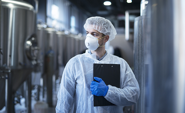 Technologist in protective white suit with hairnet and mask standing in food factory.