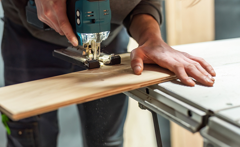 Worker saws wooden shelf on saw table with jig saw.