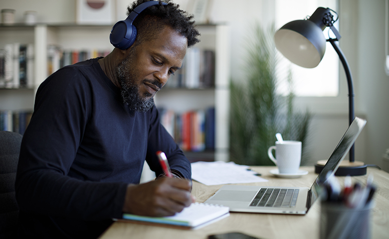 Man with headphones working on a laptop and taking notes on paper